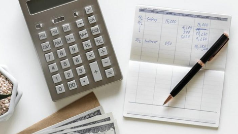 A notepad is sitting on a table with a household budget written on it, with a pen resting on top of the notepad and a calculator sitting next to them